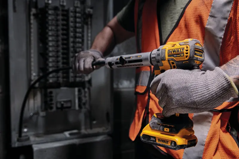 Een persoon met handschoenen en een oranje veiligheidsvest gebruikt een DEWALT 20V draadloos elektrisch gereedschap om aan een elektrisch paneel te werken.