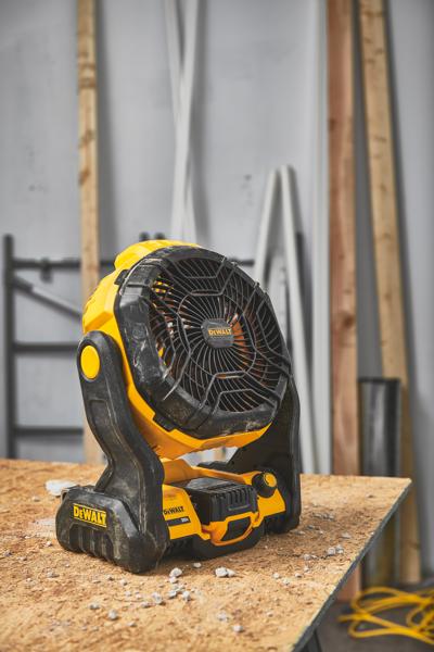 Een draagbare draadloze ventilator van DEWALT (model DCE512B) staat op een houten werkbank in een werkplaats met bouwmaterialen op de achtergrond.